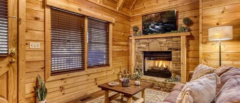 A cozy log cabin interior with a stone fireplace, wooden walls, a comfortable sofa, and a small table with decorative items.