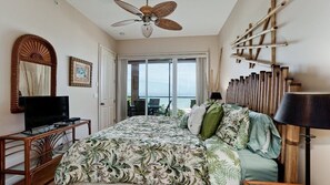 A bedroom with a bed, a television on a stand, a ceiling fan, and a view of the ocean through large windows. - Bradenton Beach Club A - Anna Maria Island- Condo 3 Bedroom/ 2 Bath ocean view, maximum occupancy of 6 people. (Bradenton Beach)