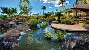 A landscaped garden with a koi pond, palm trees, and a building with a covered patio area. - Summer Sale Rates- 2BR/2BA on Sunset Side of BT (Kapolei)