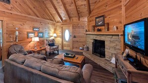 A cozy log cabin interior with a stone fireplace, a television, a sofa, and a patterned armchair.