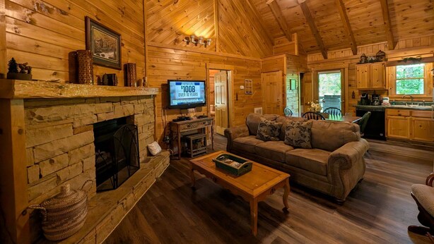A cozy log cabin interior with a fireplace, a television, a sofa, and a kitchen area.