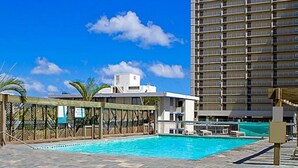 A swimming pool area with a clear blue sky, a tall building in the background, and a fenced structure with a sign. - Clean studio at the heart of Waikiki!! (Honolulu)
