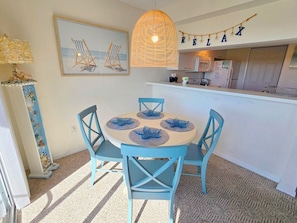 Een eetgedeelte met een ronde tafel, vier blauwe stoelen en een geweven hanglamp. Op de achtergrond is een keuken zichtbaar, met een 'RELAX' bord boven de toonbank.