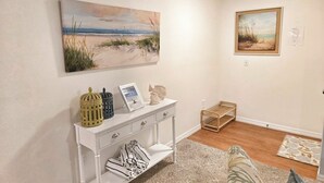 Une pièce comprenant une console blanche, un tableau représentant une scène de plage, un tableau encadré et un petit panier en bois.