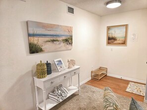 Een kamer met een witte console tafel, een schilderij van een strandlandschap, een lijst met een foto en een klein houten mandje.