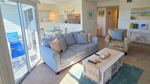 Un salon avec un canapé, un fauteuil et une table basse, une cuisine à l’arrière-plan et une porte-fenêtre coulissante donnant sur un espace extérieur.