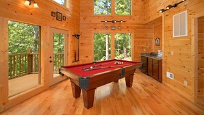Interior de una cabaña de madera con mesa de billar, zona de cocina y amplios ventanales con vistas al bosque.