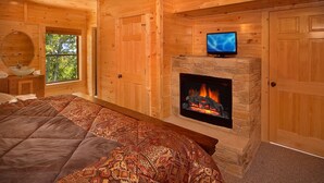 Habitación tipo cabaña de madera con chimenea de piedra, un televisor montado encima y una cama grande con una manta estampada.