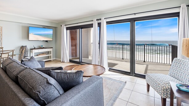 Een woonkamer met een grote televisie, grijze banken, een houten salontafel en een balkon met uitzicht op het strand.