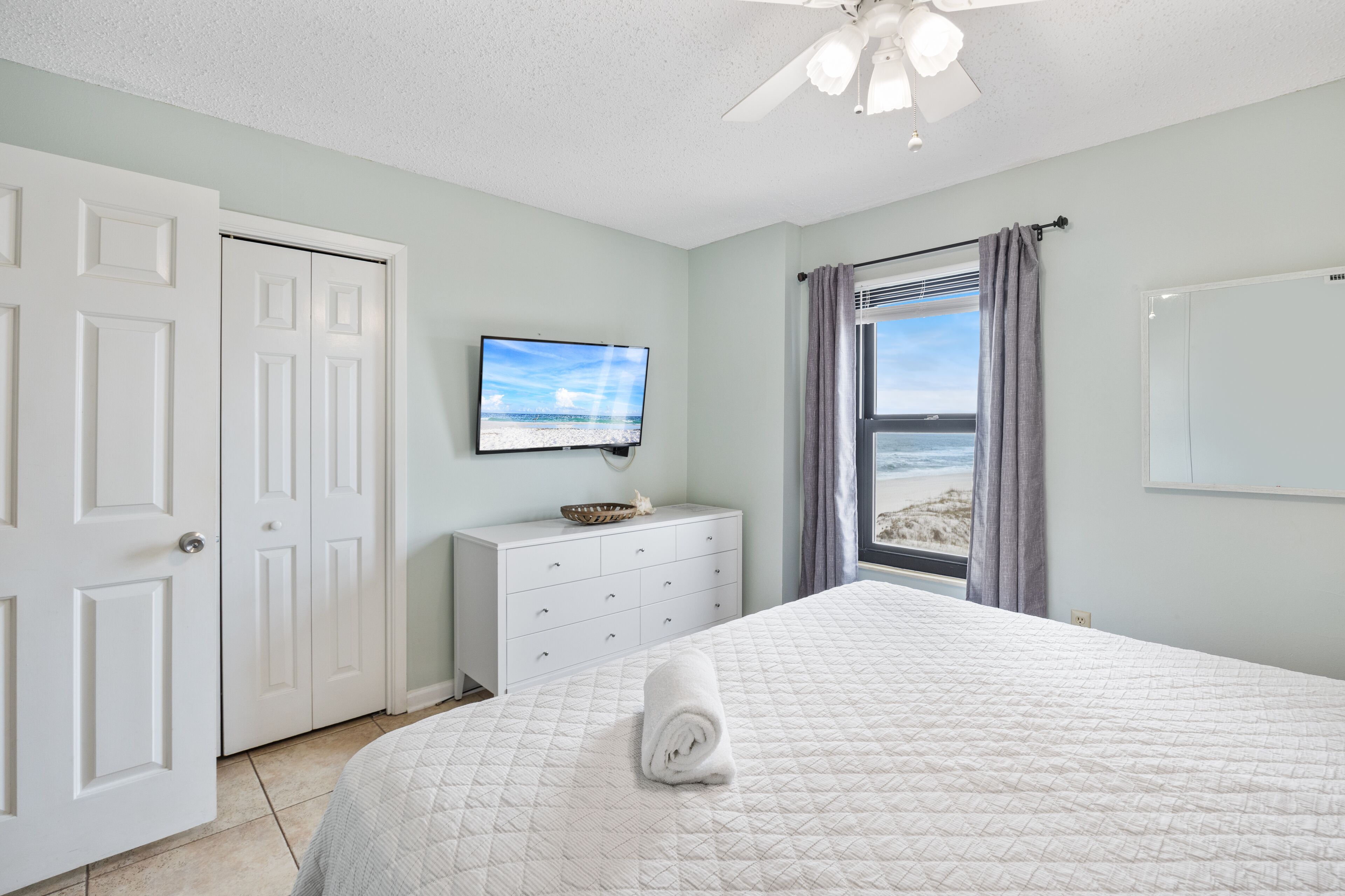 Un dormitorio con cama, tocador, televisor y ventana con vista a la playa.
