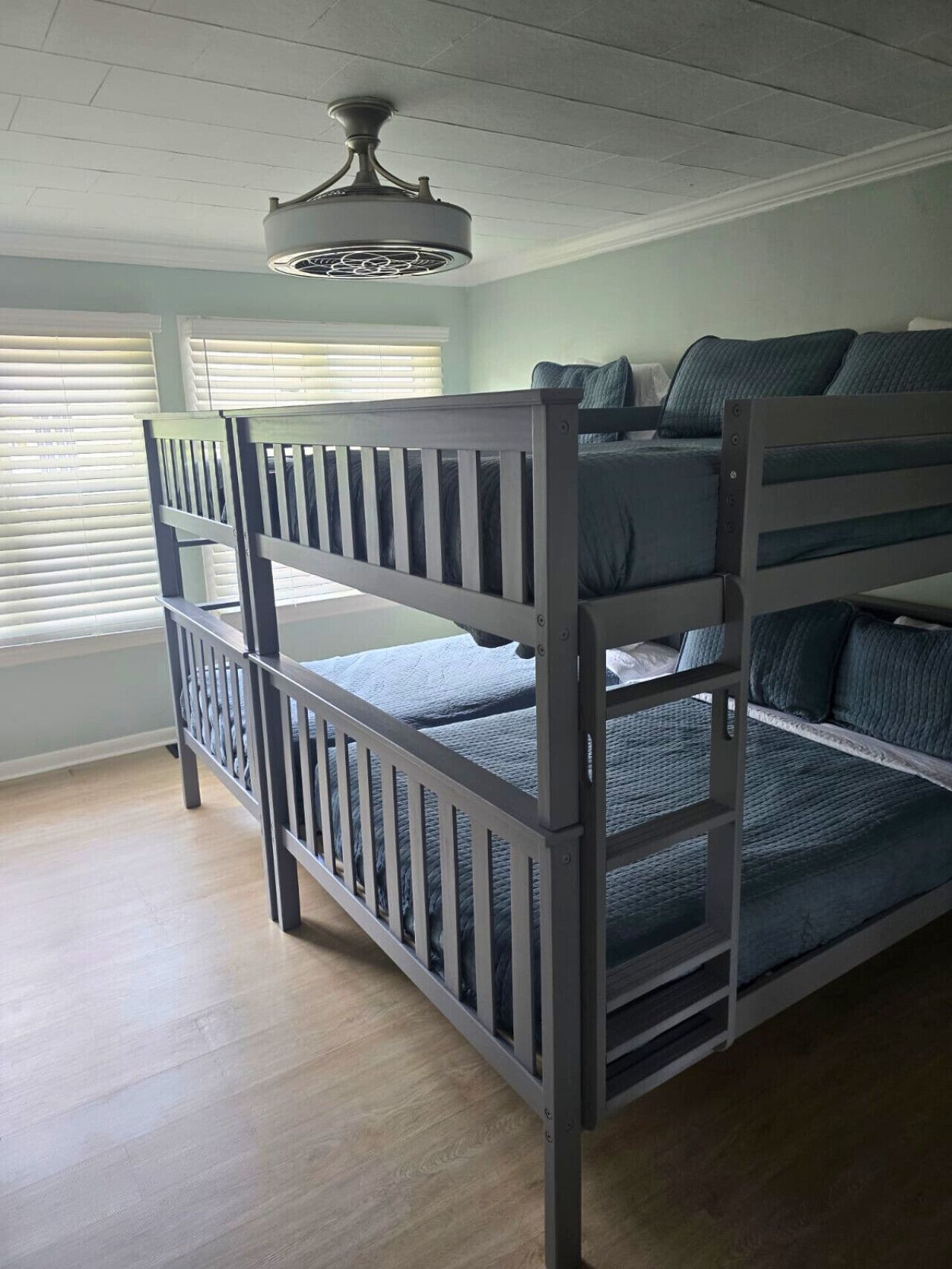 A bunk bed with a ceiling fan, light-colored walls, and a window with blinds.
