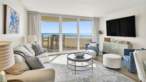 Ein geräumiges Wohnzimmer mit großem Fenster zum Meer, einem wandmontierten Flachbildfernseher und einer gemütlichen Sitzecke mit Sofa und Hocker.