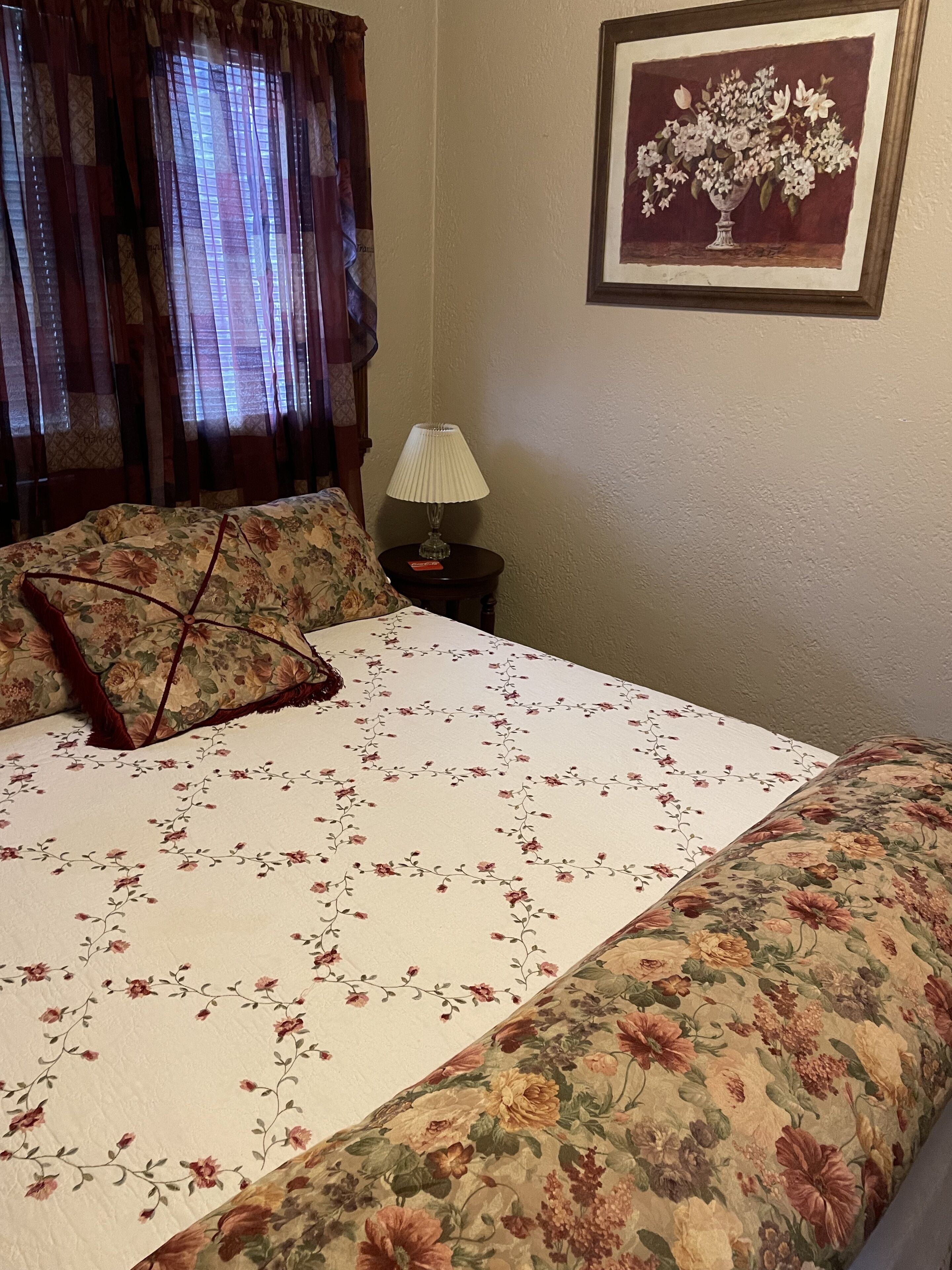 A neatly made bed with floral bedding, a bedside lamp, and a framed floral artwork on the wall.