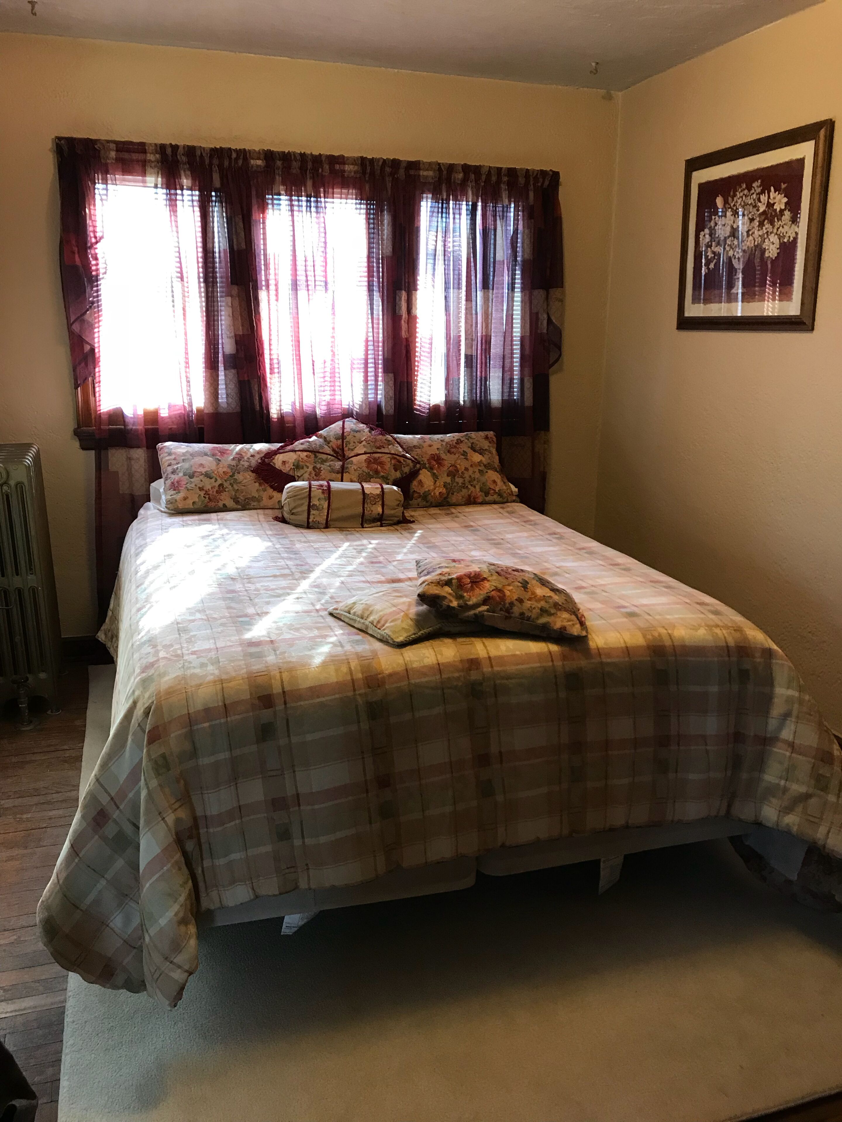 A bedroom with a bed, floral-patterned pillows, a plaid bedspread, and a window with curtains.