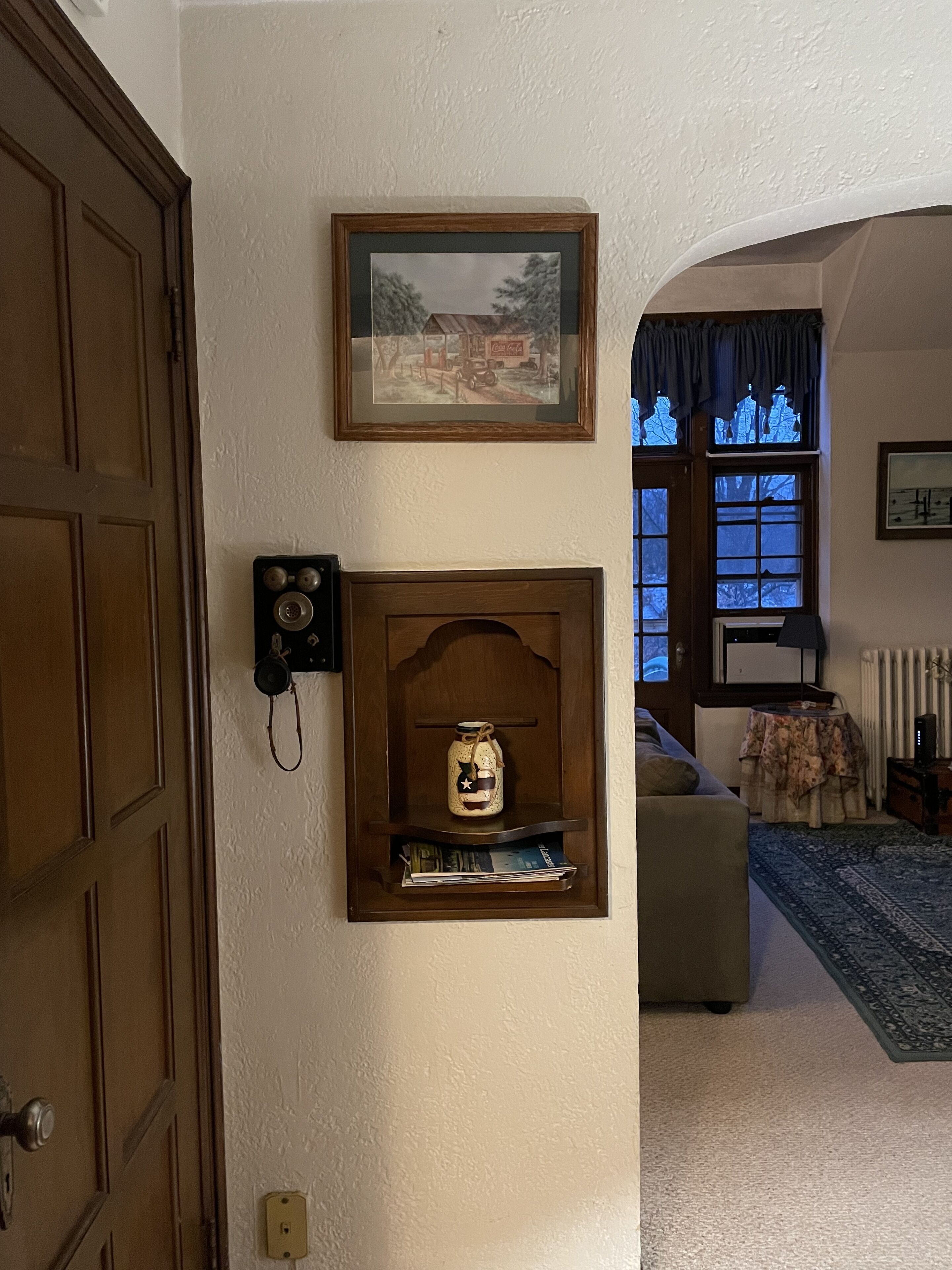 Ein Zimmer mit Holztür, einem gerahmten Bild an der Wand, einem Regal mit einer Dekoration, einem Fenster mit blauen Vorhängen und einer Heizung.