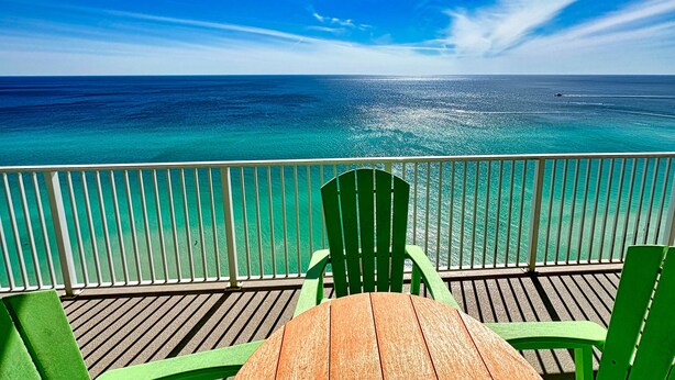 Een houten terras met een tafel en groene stoelen, met uitzicht op een turquoise oceaan en een heldere hemel.