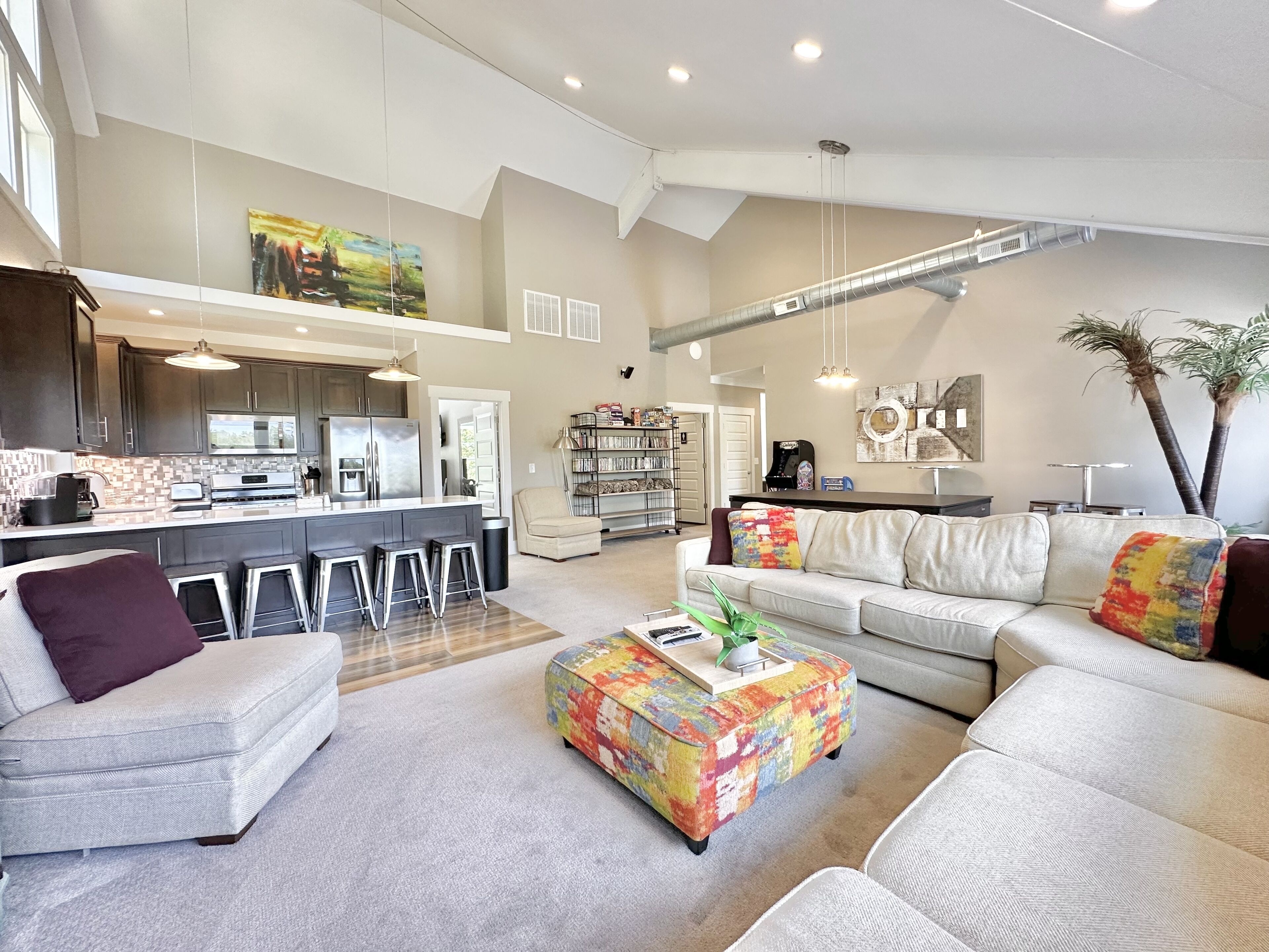 A spacious living area with a large sofa, a colorful ottoman, and a kitchen in the background.