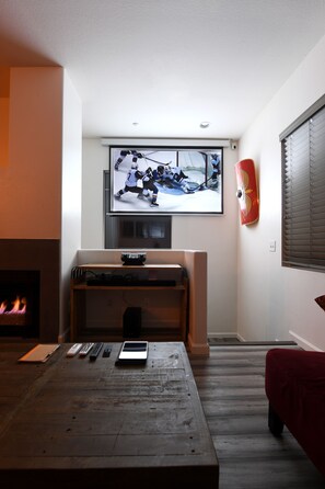 A living room with a TV mounted on the wall, a fireplace, and a wooden coffee table with remote controls.