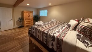 A bedroom with a wooden bed frame, a large wardrobe, a standing fan, and a window with blinds.