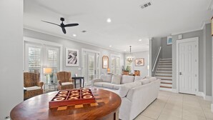 A spacious living room with a round wooden table, a grey sofa, wicker chairs, and a staircase leading to another level. - Forever Summer: Discover Your Happy Place! (Rosemary Beach)