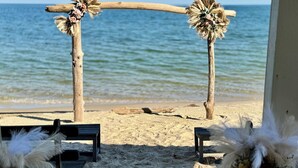 Un arco de madera con adornos florales sobre una playa arenosa, que conduce a una zona con bancos y arreglos de plumas blancas.