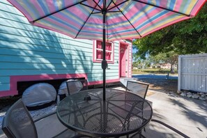 A patio with a round metal table and chairs, a colorful striped umbrella, and a house with blue siding and pink accents.