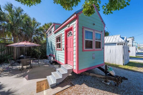 A small, single-story house with a pink door and windows, a porch with a red umbrella, and a gravel driveway.