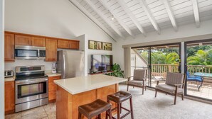 Una cocina con armarios de madera, electrodomésticos de acero inoxidable, una isla grande con taburetes y un ventanal corredizo que da a un balcón con mobiliario exterior.