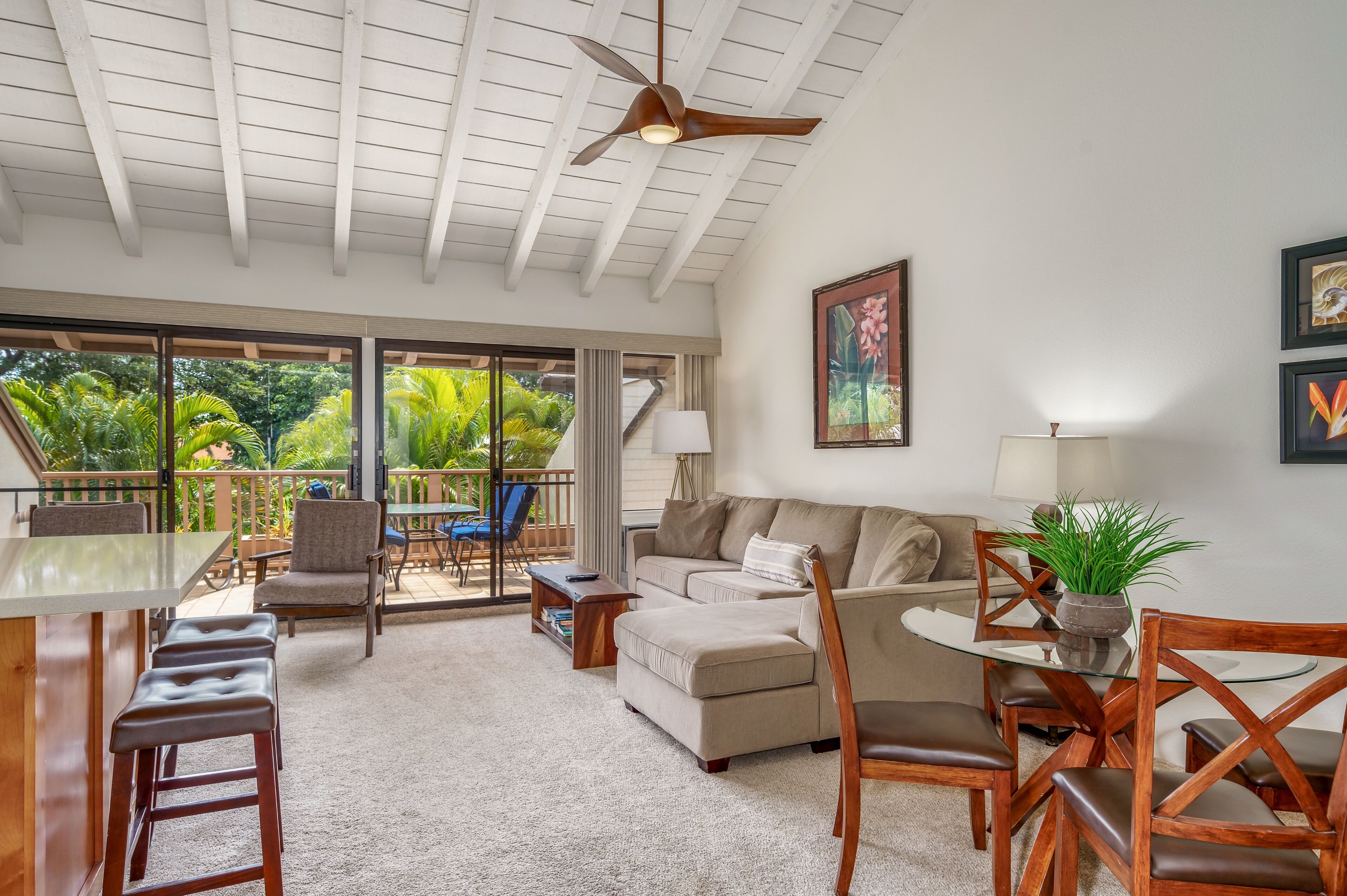 A spacious living area with a ceiling fan, a sofa, a coffee table, and a dining table with chairs. There is a balcony with a view of greenery.