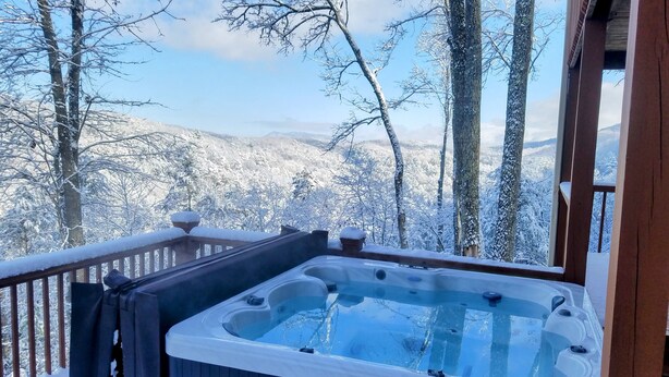 A hot tub on a wooden deck with snow-covered trees and mountains in the background.