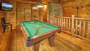 Ein Holzbilliardtisch mit Kugeln, ein Fernseher und eine Steinsäule in einem Blockhausszimmer.