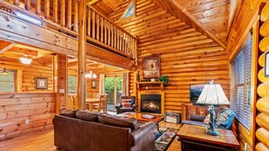 Un interior acogedor de cabaña de madera con chimenea, sofá de cuero, pisos de madera y un balcón.