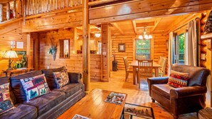 Interior de una cabaña de troncos con una escalera de madera, un comedor y una sala de estar con sofás de cuero y una mesa de centro.