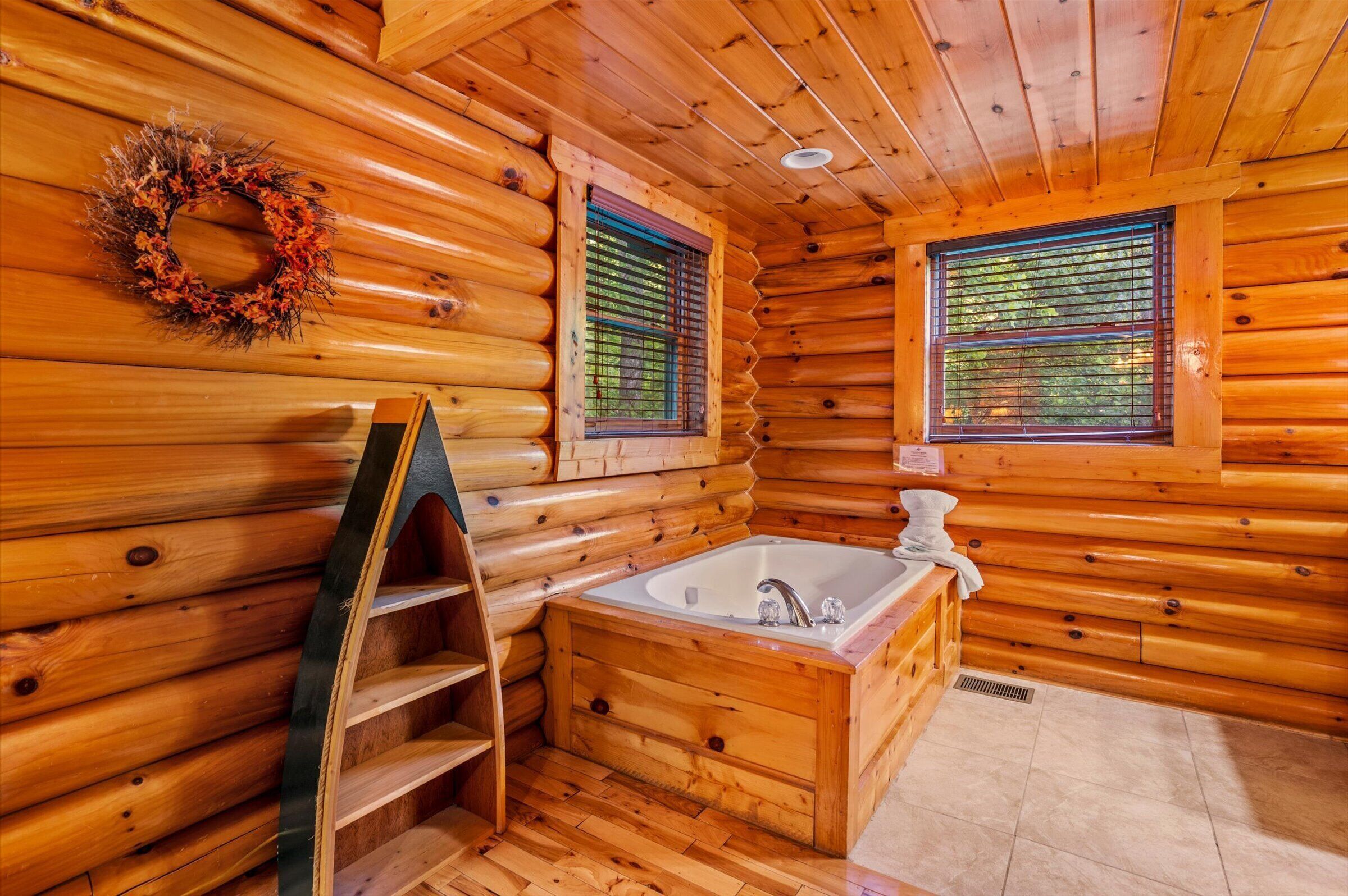 A log cabin bathroom with a wooden bathtub, two windows with blinds, a towel rack, and a wreath on the wall.