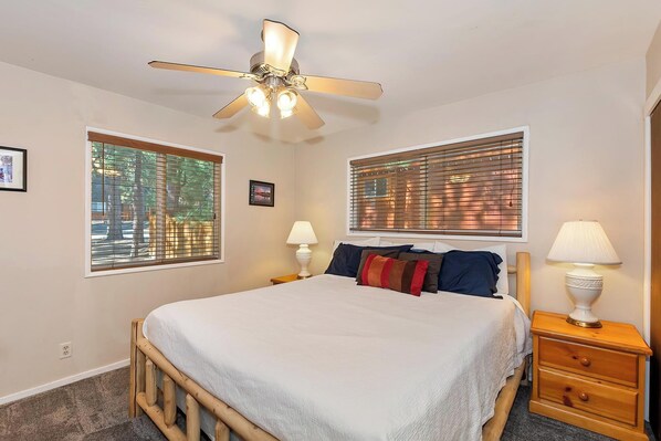 A bedroom with a bed, two bedside tables with lamps, a ceiling fan, and two windows with blinds.