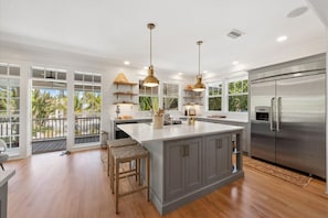 A spacious kitchen with a large island, stainless steel appliances, and a view of palm trees through large windows.