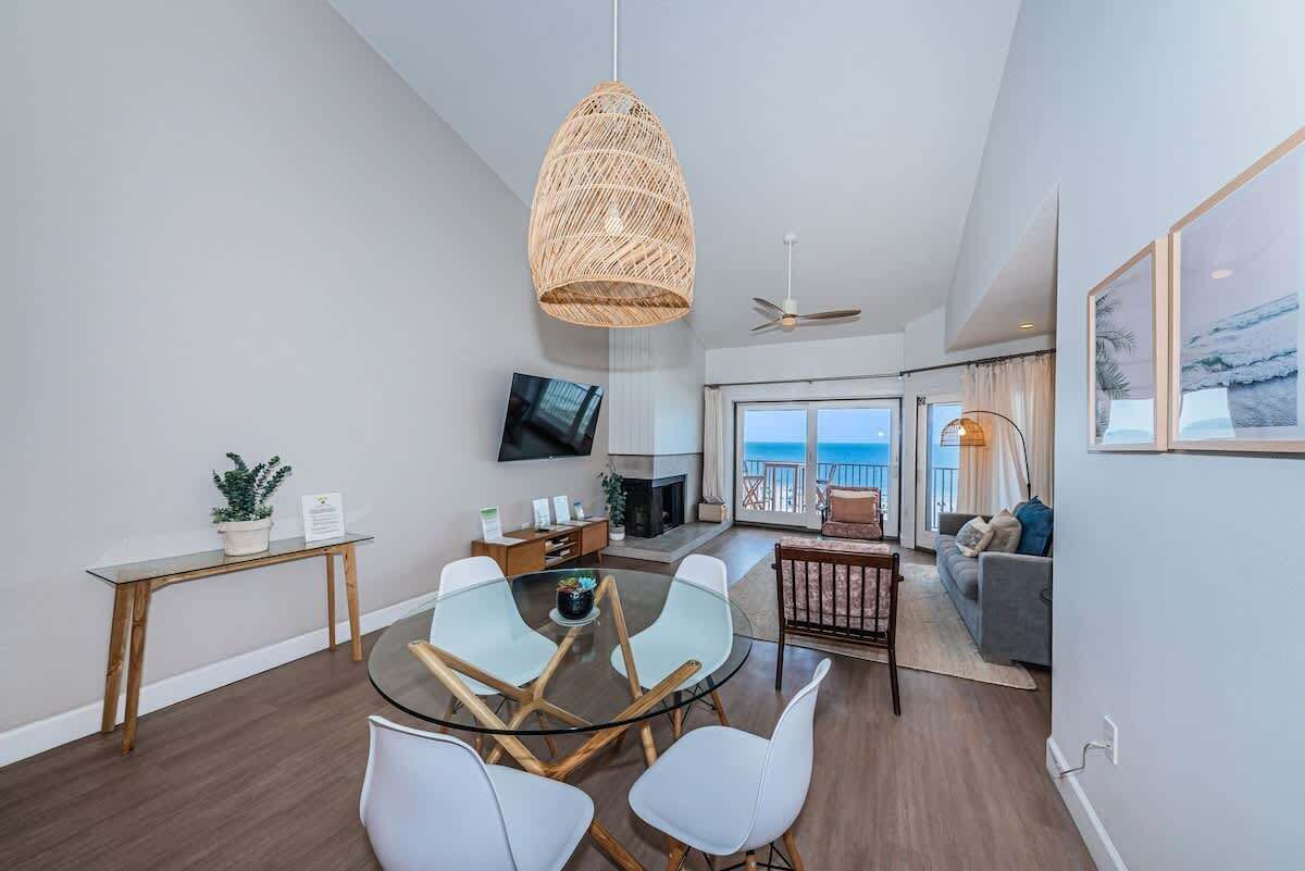 A modern living room with a glass dining table, white chairs, a television, and a fireplace.