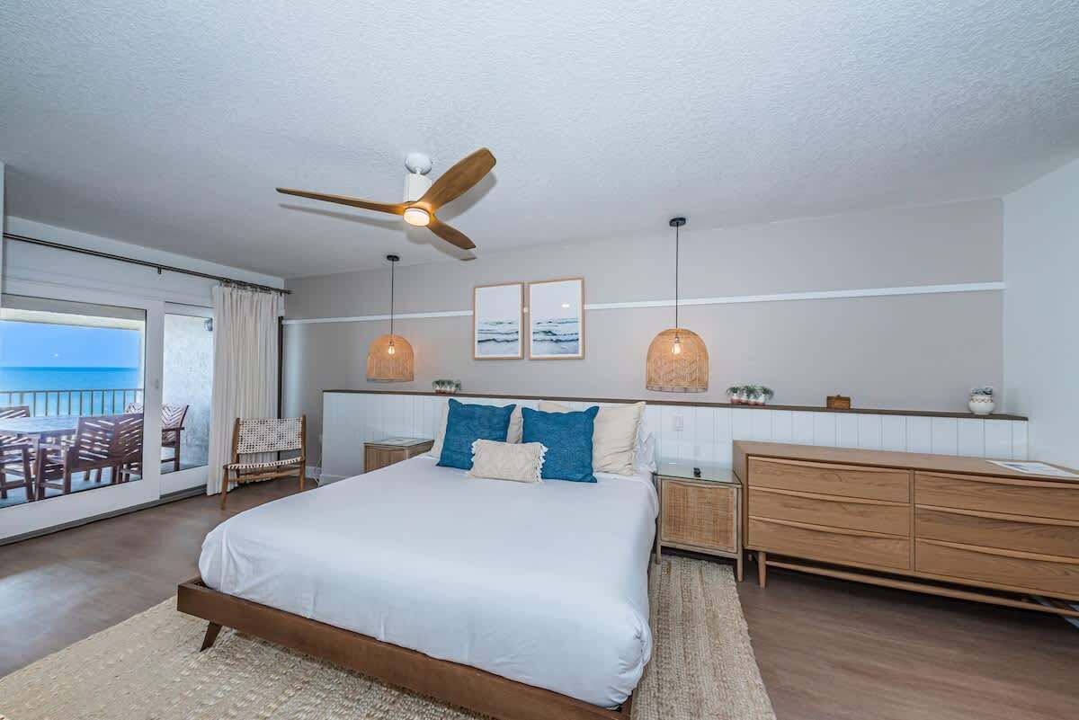 A bedroom with a bed, a ceiling fan, a dresser, a nightstand, and a view of the ocean through a sliding door.