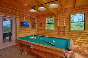 A wooden pool table in a cabin with a view of the outdoors through windows.