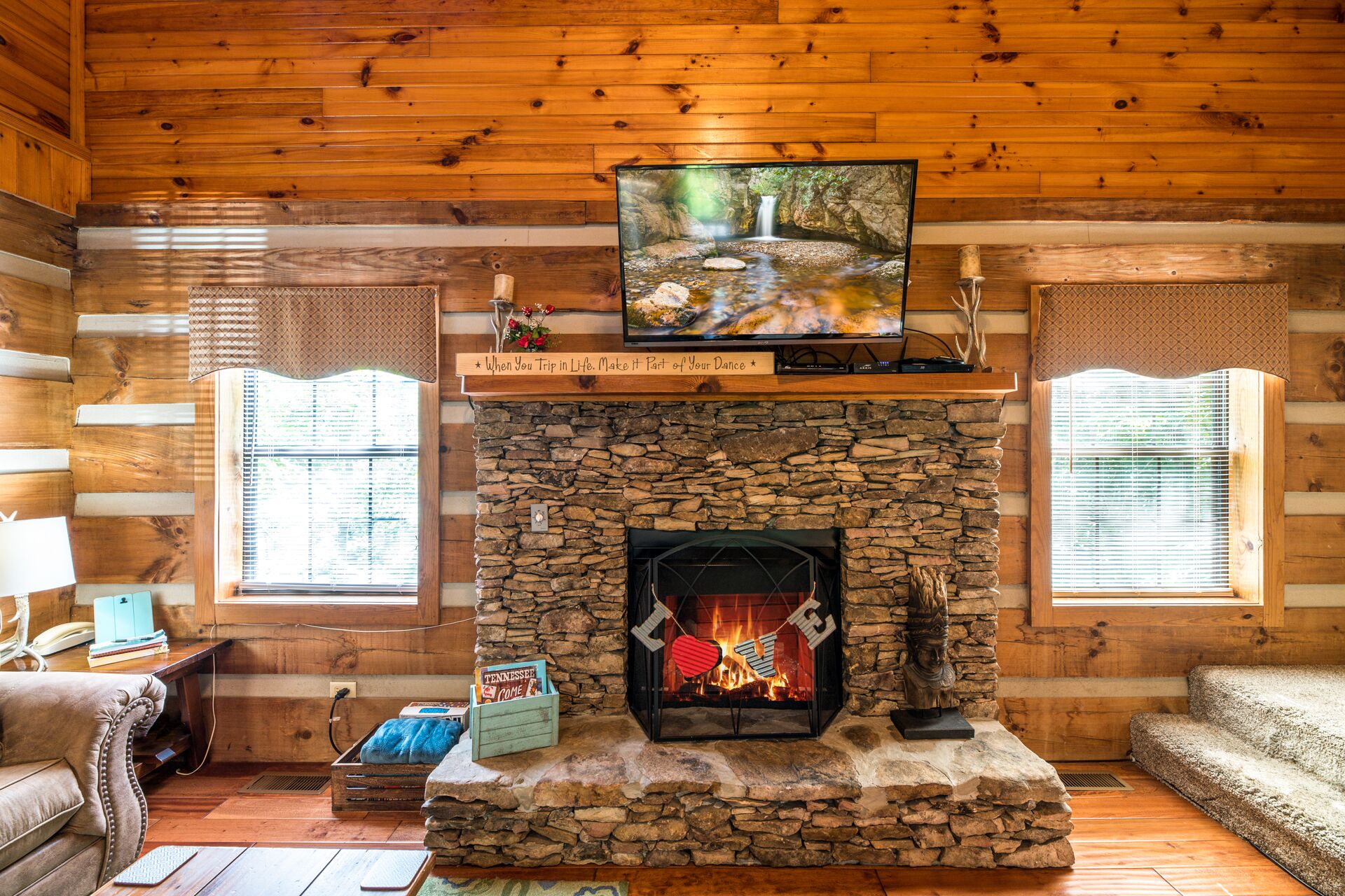 A log cabin interior with a stone fireplace, a television mounted above it, and a cozy seating area.