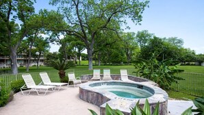 A hot tub surrounded by lounge chairs, a pool, and a fenced area with trees. - WW C301 Urban Bungalow (New Braunfels)