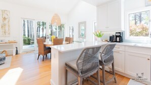 A bright kitchen with white cabinets, a central island with bar stools, and large windows offering views of palm trees and a balcony.