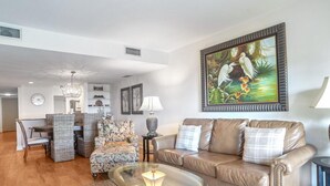 A living room with a glass coffee table, a brown leather sofa, wicker chairs, and a dining area with a clock and shelves. - Ocean One 315 - Oceanfront 3rd Floor Condo (Hilton Head Island)