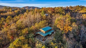 A two-story log cabin with a green roof, surrounded by dense forest with autumn foliage. - Take My Breath Away: Mountain Views, Theater (Gatlinburg)
