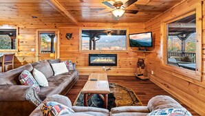 A cozy cabin interior with a fireplace, wooden walls, a ceiling fan, and large windows offering views of the outdoors. - Take My Breath Away: Mountain Views, Theater (Gatlinburg)