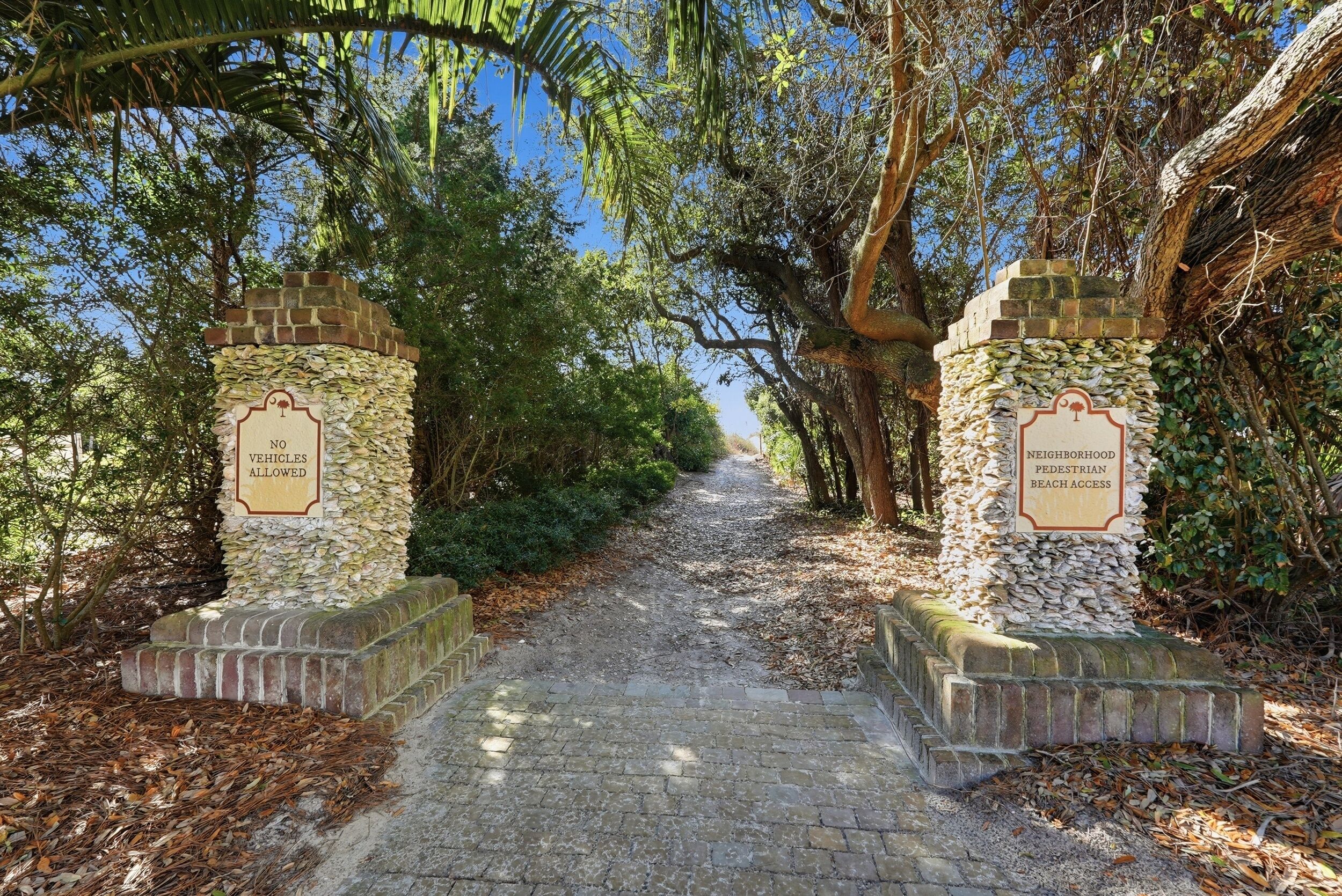 A stone pathway leading to a beach, flanked by stone pillars with signs indicating no vehicles allowed and neighborhood pedestrian beach access.