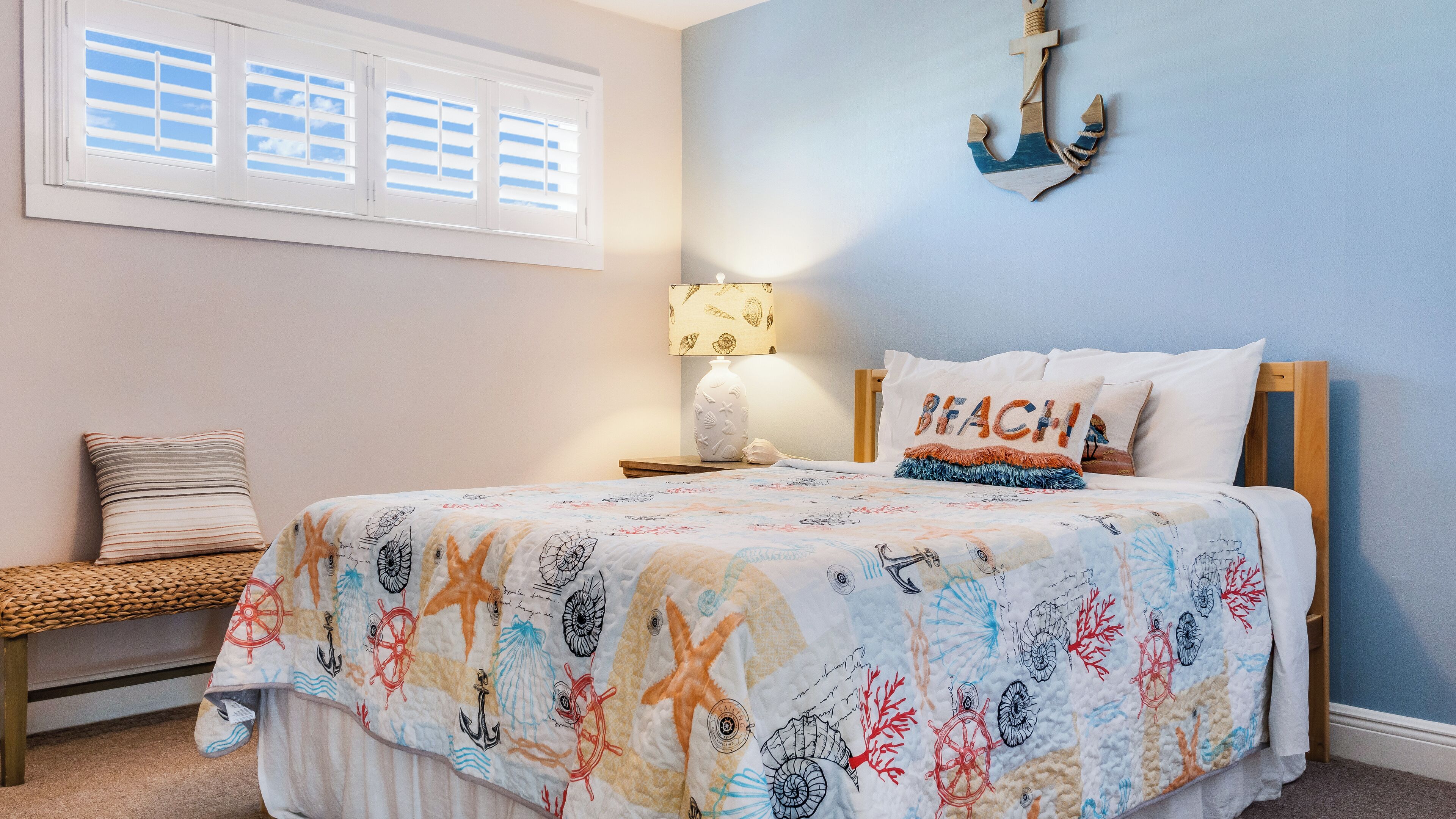 A bedroom with a bed, a nightstand, a lamp, a window with blinds, and a wall-mounted anchor decoration.