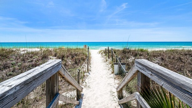 Ein hölzerner Steg führt zu einem Strand mit klarem, blauem Wasser und sandigem Ufer.