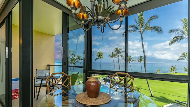 A glass table with a vase, chairs, and a view of the ocean and palm trees. - Oceanfront Retreat | Split A/C | Pool | Walk to Beach & Dining (KIHEI)