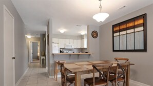 Un coin repas avec une table et des chaises en bois, une cuisine à l’arrière-plan et une horloge au mur.